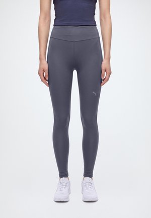 Un mannequin porte un legging Puma gris foncé à taille haute et des baskets blanches, debout devant un fond uni.