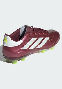 Botín de fútbol color marrón con diseño de tres rayas blancas, presenta una textura superior suave y tachuelas de color verde brillante para tracción en las superficies del campo.