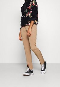 Felpa nera con motivo floreale, pantaloni beige slim-fit con fessure laterali e sneakers alte nere con suola bianca, presentati su uno sfondo semplice.