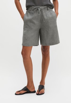 Osoba nosząca szare lniane szorty ze sznurkiem, czarny dzianinowy top i czarne skórzane sandały z odkrytymi palcami, stojąca z rękami w kieszeniach.