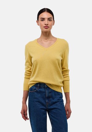 Femme aux cheveux bruns tirés en arrière, portant un pull jaune à col en V et un jean bleu taille haute, devant un fond clair.