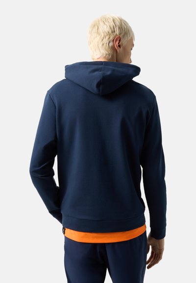 Blond osoba v námořnicky modré mikině přes oranžové tričko, otočená zády, stojí před jednoduchým světlým pozadím.