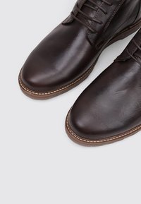 Botas de cuero marrón hasta el tobillo con un acabado suave, con diseño de cordones, punta redonda y costuras beige contrastantes a lo largo de la suela.