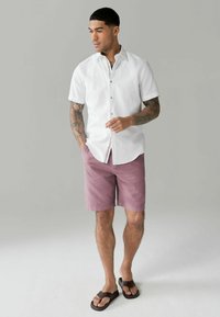 Hvid kortærmet skjorte med knapper, skræddersyet fit, parret med lyserøde shorts og brune flip-flops, mod en neutral baggrund.