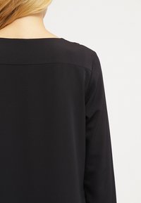 Haut noir à manches longues avec un décolleté carré, texture lisse et design minimaliste, sans quincaillerie ni motifs visibles.