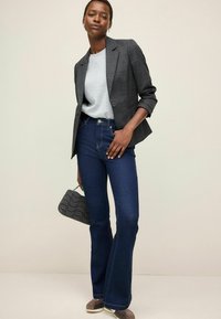 Blazer a quadri grigio, t-shirt grigia, jeans a campana blu scuro, borsa grigia con texture, scarpe marroni, in piedi su uno sfondo chiaro.