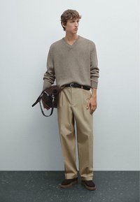 Sweter w serek w kolorze taupe, zestawiony z beżowymi spodniami w fałdy. Ciemnobrązowe zamszowe buty i brązowa torba na ramię z paskiem.
