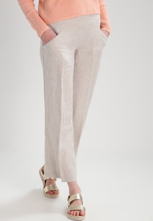 Persoon die lichtbeige wijde broek met zakken draagt, een perzikkleurige gebreide trui en gouden sandalen met open tenen, staand tegen een effen achtergrond.