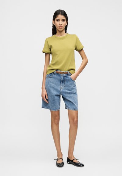 Λαδί κοντομάνικο μπλουζάκι με στρογγυλή λαιμόκοψη, συνδυασμένο με ανοιχτό μπλε τζιν σορτς και μαύρα παπούτσια. Απλός σχεδιασμός με casual εφαρμογή.