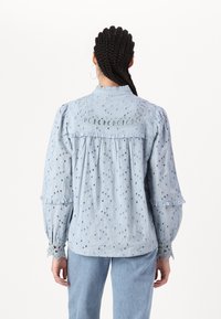 Light blue blouse made of broderie anglaise fabric, featuring puffy sleeves, ruffled accents, and cut-out details along the yoke and cuffs.