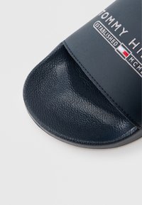 Tumši zils slaids sandāls ar teksturētu zolīti, gludu sintētisku siksniņu, uz kuras ir balts "TOMMY HILFIGER" logotips un dibināšanas gads kontrastējošās krāsās.