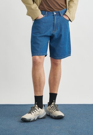 Muž oblečený v modrých rifľových šortkách, béžovej bunde, hnedom tričku, čiernych ponožkách a béžových turistických topánkach stojí na modrom koberci.