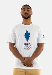 Camiseta de algodón blanca con un gráfico de llama azul, la palabra "FRANCIA" y cinco anillas olímpicas de colores en el frente. Manga corta.