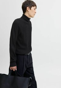 Suéter negro de cuello alto acanalado combinado con pantalones negros; sosteniendo un gran bolso de cuero negro. Textura suave, diseño ajustado.