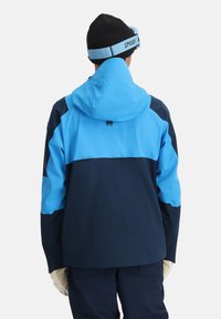 Blau- und marineblaue isolierte Jacke mit Kapuze, mit einem Farbblockdesign und elastischen Bündchen, getragen mit beigen Handschuhen und einer schwarzen Mütze.