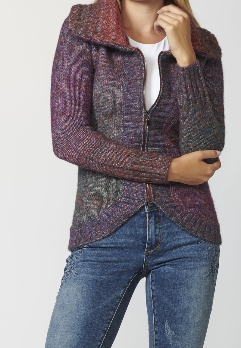 Femme portant un pull zippé en tricot multicolore avec un grand col par-dessus une chemise blanche, assorti à un jean bleu brodé.