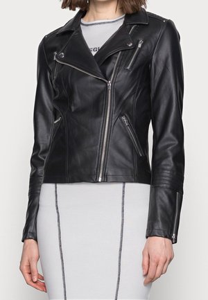 Femme portant une veste de motard en cuir noir ajustée avec des fermetures éclair argentées sur une robe gris clair avec des coutures verticales foncées.