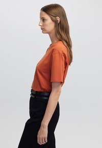 T-shirt arancione a maniche corte con scollo rotondo, realizzata in un tessuto liscio. Abbinato a pantaloni neri a vita alta e una sottile cintura nera.