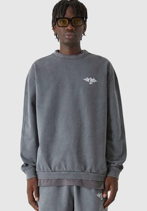 Sweatshirt gris surdimensionné en tissu doux avec un col rond, des poignets côtelés et un petit logo blanc sur la poitrine.