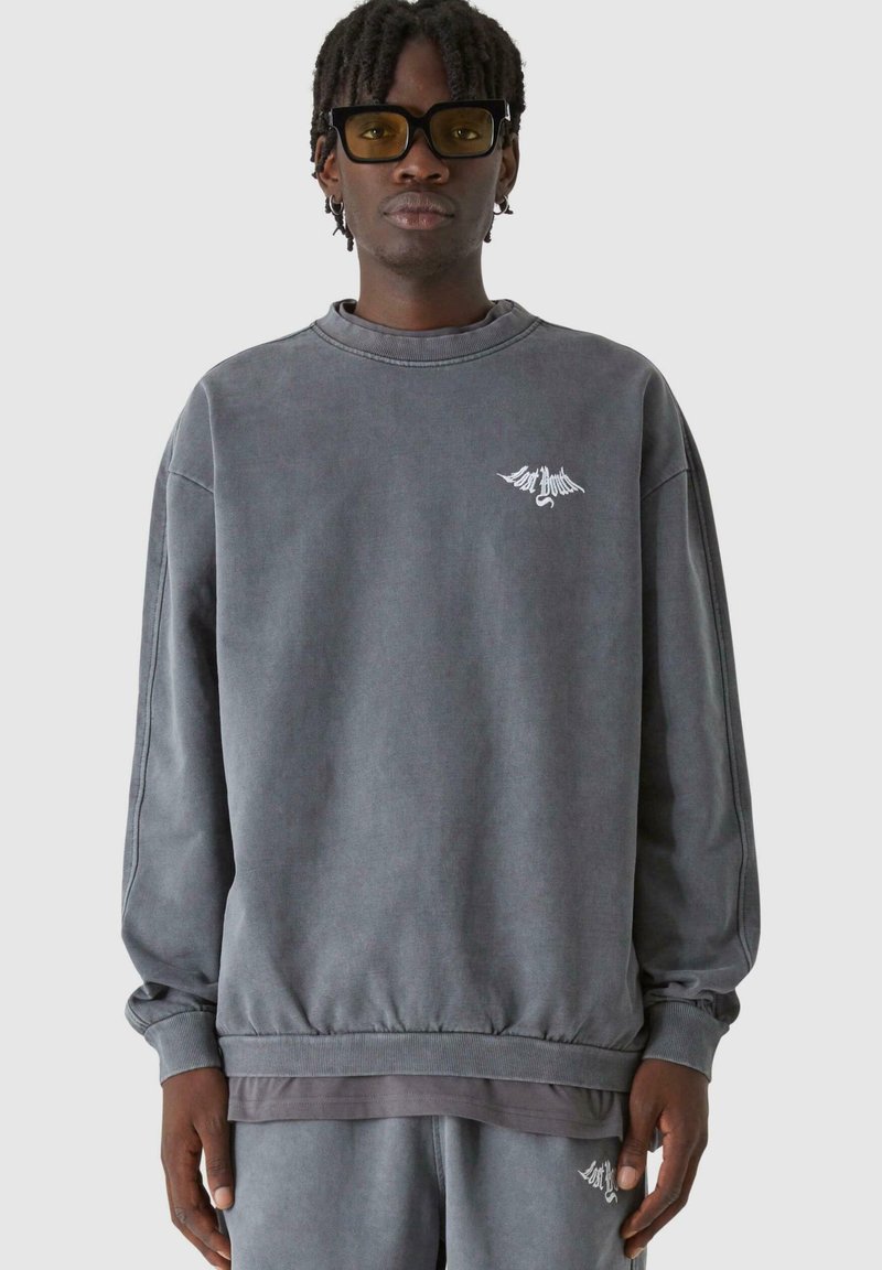 Szürke, oversize pulóver, puha anyagból, kerek nyakkal és bordázott mandzsettával, a mellkason pedig egy apró fehér logó található.