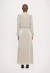 Robe longue beige clair avec une texture brillante, manches longues et jupe à volants. Comprend une ceinture en chaîne argentée à la taille.