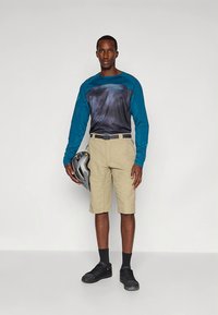 Långärmad skjorta i en gradient från teal till marinblå, tillsammans med beiga shorts. Modellen håller i en grå hjälm och bär svarta sneakers och strumpor.