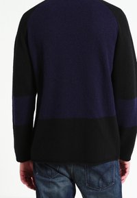 Man i långärmad tröja med svarta och mörkblå block och blå denimjeans, vänd bort från kameran.