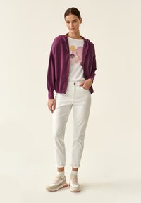 Cardigan lavorato a maglia viola con cappuccio, indossato sopra una maglietta bianca con motivi astratti colorati, abbinato a jeans bianchi a gamba stretta e sneakers.