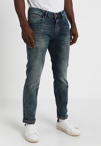 Jeans de mezclilla azul con un ligero desgaste, cinco bolsillos y una etiqueta de marca en la parte trasera. Llevados con una camiseta blanca y zapatillas blancas.