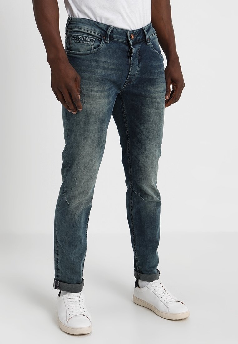 Jeans de mezclilla azul con un ligero desgaste, cinco bolsillos y una etiqueta de marca en la parte trasera. Llevados con una camiseta blanca y zapatillas blancas.