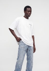 Jeune homme portant un t-shirt blanc ample et un jean déchiré bleu clair en patchwork, debout devant un fond blanc uni.