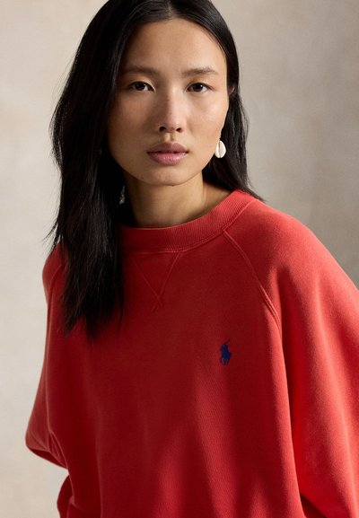 Γυναίκα με μακριά μαύρα μαλλιά που φοράει κόκκινο φούτερ με μικρό μπλε κεντημένο λογότυπο και λευκά σκουλαρίκια σε σχήμα κοχυλιού, ουδέτερο φόντο.