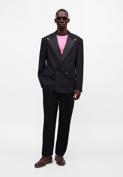 Homme portant un blazer noir croisé avec des revers en satin, une chemise rose, un pantalon noir, des chaussures marron et des lunettes de soleil sur fond blanc.