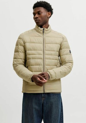 Jeune homme aux cheveux courts et bouclés, portant une veste matelassée beige et un jean bleu, debout, les mains jointes, regardant sur le côté.