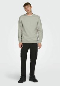 Světlě šedá mikina s žebrovanými manžetami a límečkem, doplněná černými cargo kalhotami s bočními kapsami a texturovanou látkou. Černé tenisky.