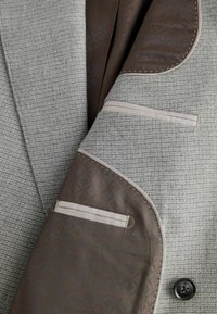 Grau gemusterter Blazer mit brauner Innenausstattung und geometrischem Design. Beinhaltet beigefarbene Akzente und einen schwarzen Knopf an der Vorderseite.