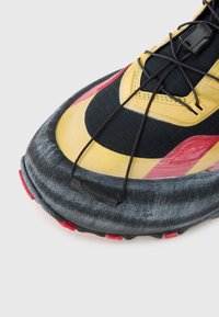 Μαύρο και κίτρινο αθλητικό παπούτσι με υφή επιφάνεια, καμπυλωτό σχέδιο και κόκκινες λεπτομέρειες. Διαθέτει μαύρ α κορδόνια και σύστημα ασφαλούς εφαρμογής.