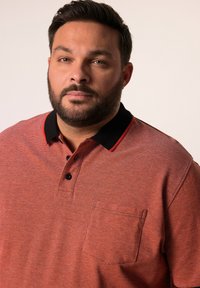 Punainen teksturoitu polo-paita, jossa on musta kaulus, jossa on punaisia yksityiskohtia ja kolme mustaa nappia. Mukana on vasemmalla rinnalla tasku.