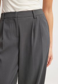 Pelēkas pludlīnijuma bikses ar gludu audumu, plakanu jostasvietu un jostu cilpām. Kreisajā pusē ir redzami sānu kabatas.