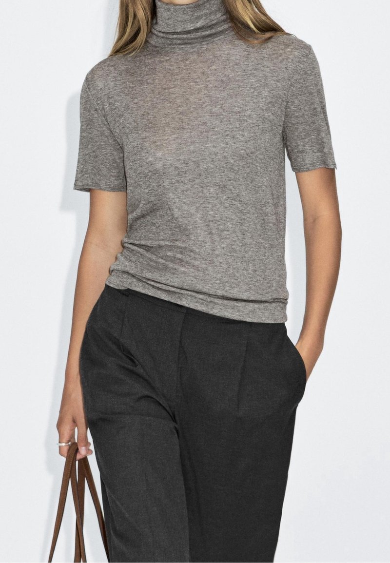 Femme portant un t-shirt gris à col roulé à manches courtes et un pantalon noir taille haute, tenant des anses marron de sac à main dans la main gauche.