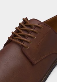 Καφέ δερμάτινο παπούτσι φόρεμα με στρογγυλή μύτη και καφέ κορδόνια που περνούν μέσα από πέντε οπές σε λείο επάνω μέρος.