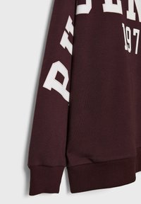 Manche de sweat-shirt marron avec lettres blanches style varsity et bord-côte, montrant une partie du texte "197" sur le corps.