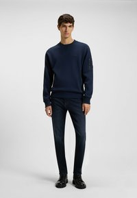 Sudadera de cuello redondo en color azul marino con tela texturizada, puños ribeteados y un bolsillo con cremallera en la manga izquierda. Combinada con jeans azul oscuro y botas negras.