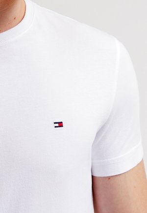 Biała bawełniana koszulka z krótkim rękawem, z małym haftowanym logo w kolorach czerwonym, niebieskim i białym na lewej stronie klatki piersiowej.