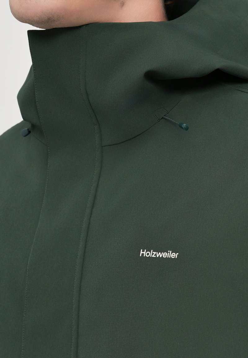Veste à capuche verte en tissu lisse, avec un col haut, des coutures minimales et un logo "Holzweiler" blanc à l'avant.