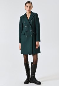 Donkergroen dubbele-breasted jas met zilverkleurige knopen, zijzakken en een verfijnde pasvorm. G gedragen over zwarte kniehoge laarzen.