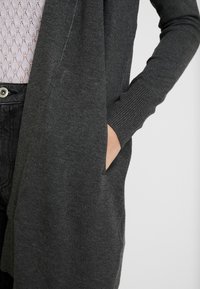 Dunkelgraue Strickjacke mit langen Ärmeln, gerippten Bündchen und zwei Seitentaschen. Weicher Stoff mit einem offenen Front-Design, über einem hellen Oberteil getragen.