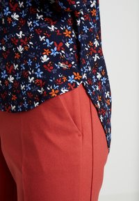 Mörkblå blus med små röda, blå och vita blommor, kombinerad med röda, texturerade byxor med sidofickor och en avslappnad passform.