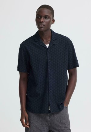 Homme portant une chemise à manches courtes, boutonnée, à motifs sombres et un jean gris, debout avec une main dans la poche, sur un fond uni.