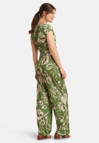 Grüner Overall mit Blumenmuster, kurzen Ärmeln, tailliert und mit weitem Bein sowie glatter Textur. Holzklotzschuhe ergänzen das Outfit.